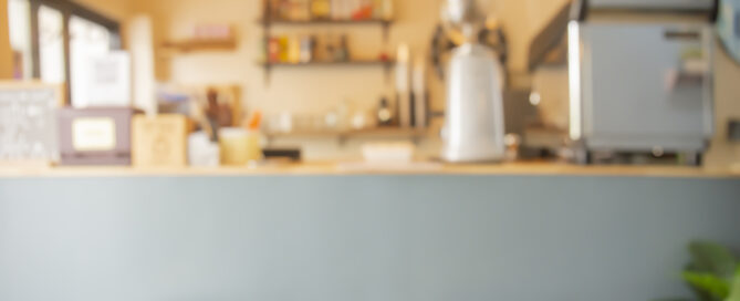 Blurred view of a small café or coffee shop counter with stools, warm lighting, and modern equipment.