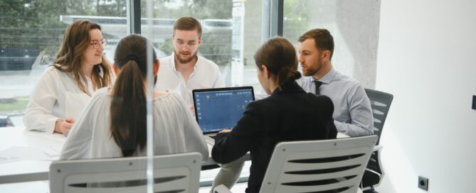 Small business team meeting in modern office discussing cash flow solutions, with laptops open on conference table and large windows in background
