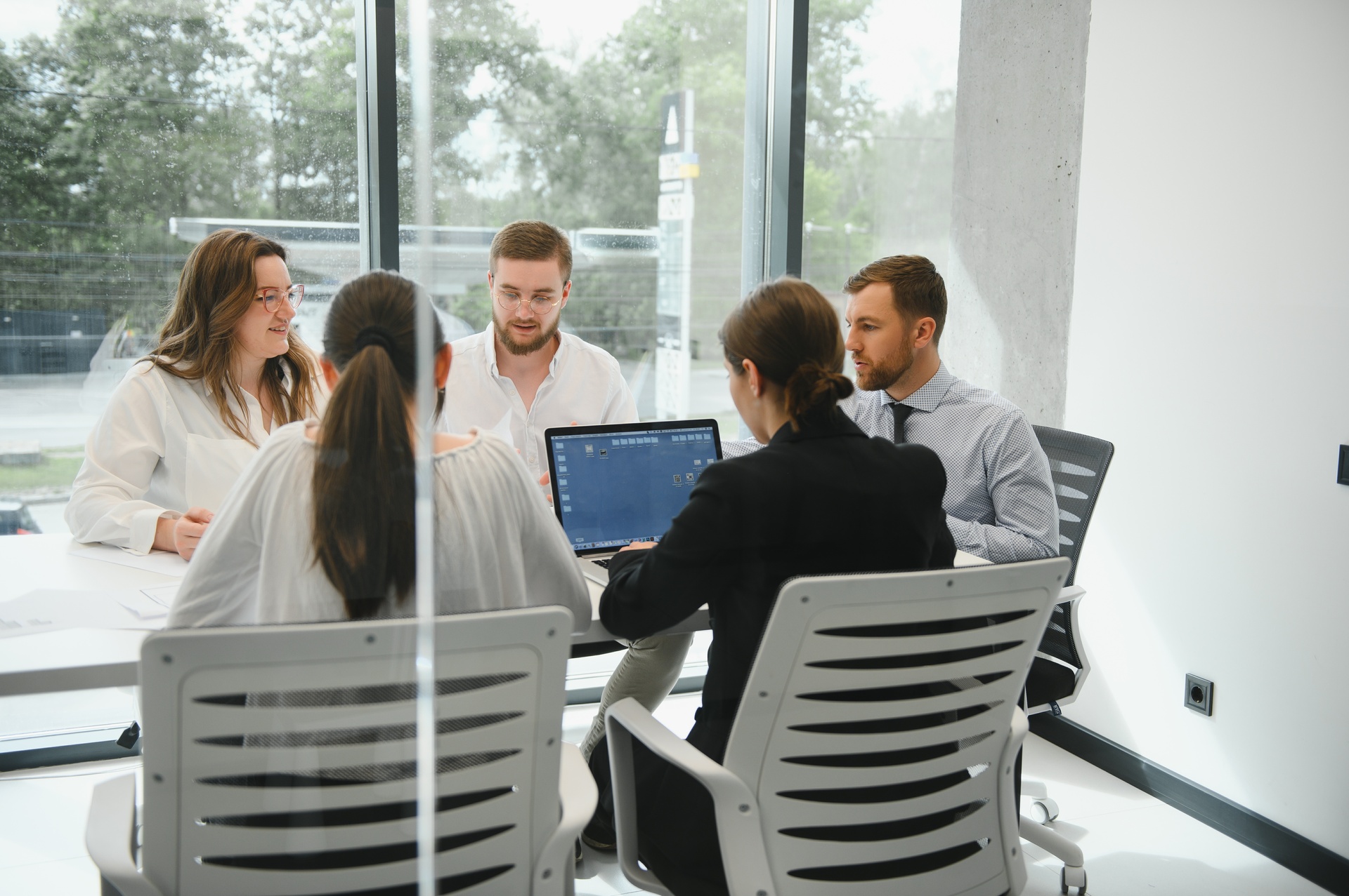 Small business team meeting in modern office discussing cash flow solutions, with laptops open on conference table and large windows in background