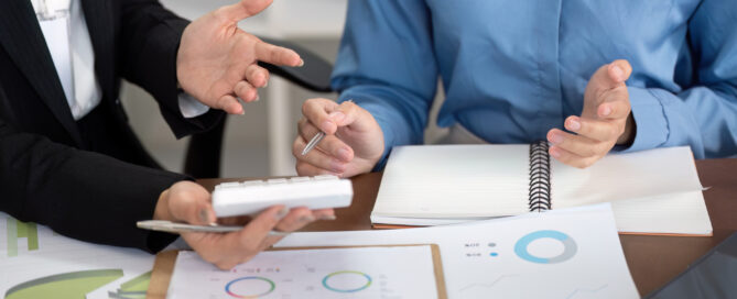 Two business professionals in consultation discussing invoice finance options, with financial charts and documents spread across a modern office desk,