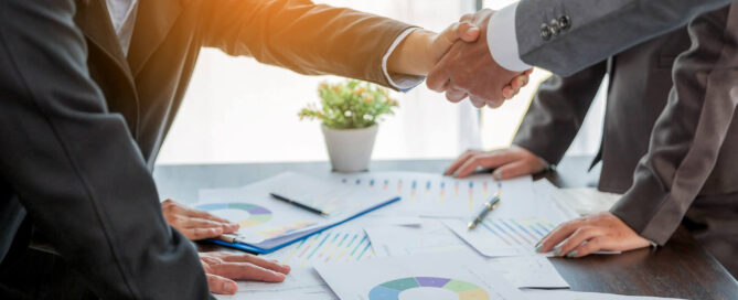 Two businesspeople in suits shaking hands across a conference table covered with charts, graphs, and financial documents in a bright office setting.