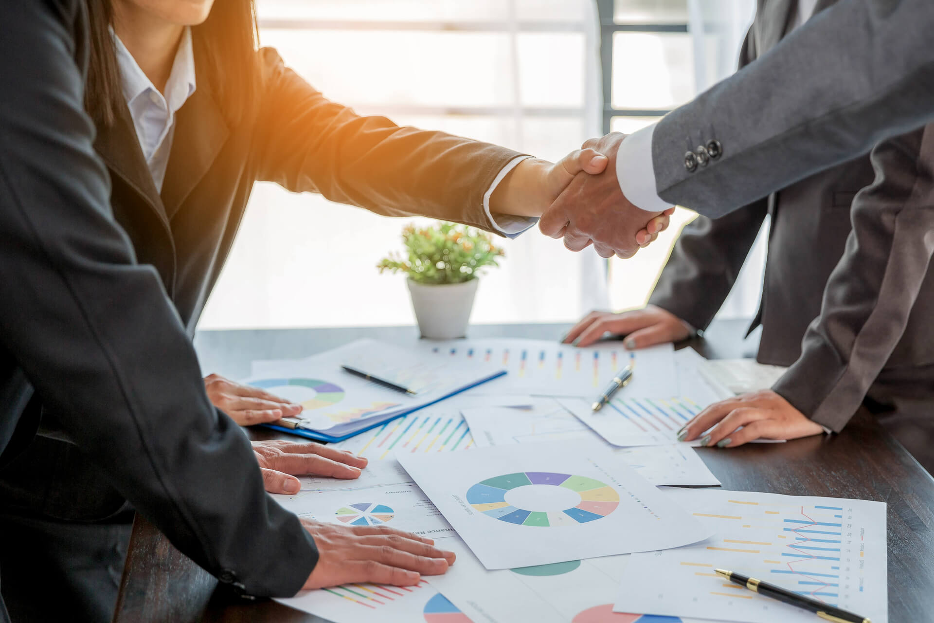 medium-vecteezy_professional-businessman-and-businesswoman-partners-shaking_10167559_medium (1) Two businesspeople in suits shaking hands across a conference table covered with charts, graphs, and financial documents in a bright office setting.