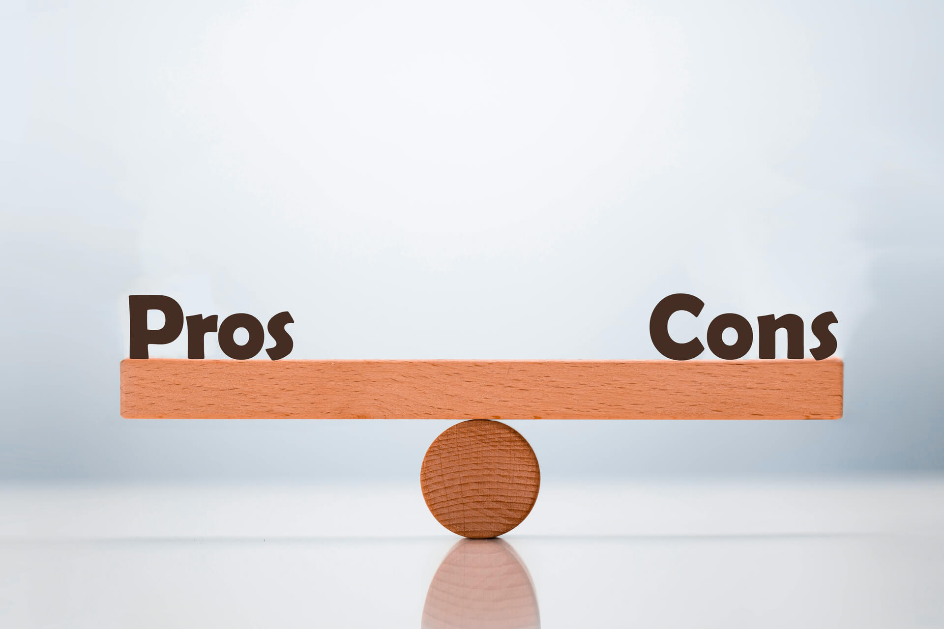 Wooden seesaw balanced on a circular fulcrum with "Pros" on the left side and "Cons" on the right side.