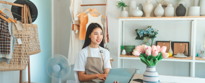 Small business owner in striped apron working on laptop in her modern retail store, surrounded by inventory and home goods, representing entrepreneurs using card revenue to fund business growth