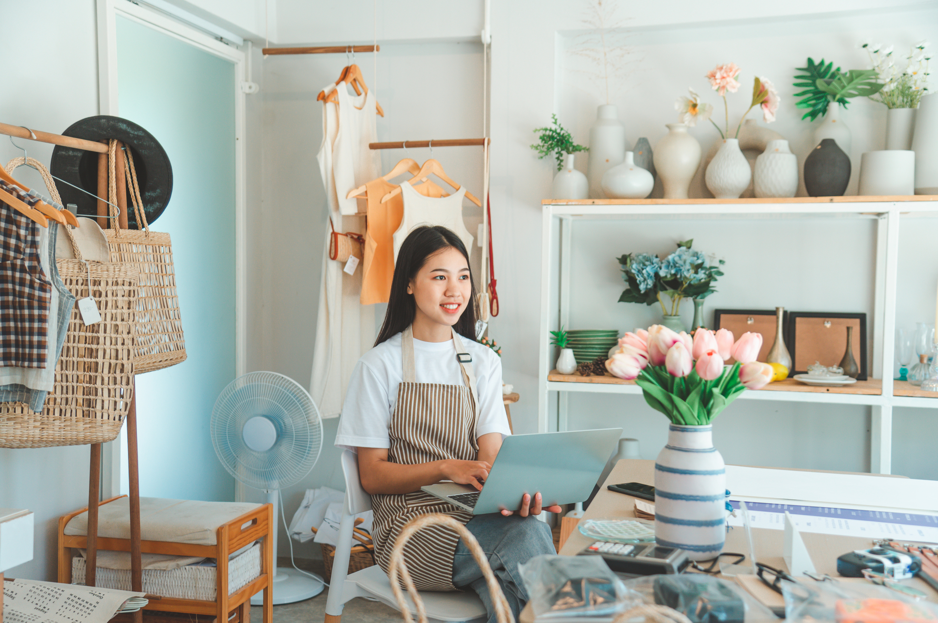 Small business owner in striped apron working on laptop in her modern retail store, surrounded by inventory and home goods, representing entrepreneurs using card revenue to fund business growth