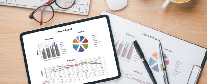 Tablet and printed finance reports with charts and graphs on a desk, surrounded by pens, glasses, keyboard, and a cup of coffee.