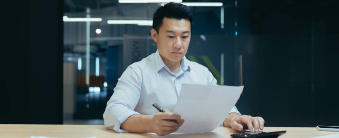 Man sitting at a desk in an office, reviewing documents while using a calculator, with papers, a tablet, and a laptop on the table.