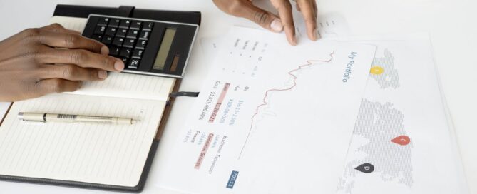 Person using a calculator with financial documents and a portfolio chart on a desk, representing business planning and financial analysis.