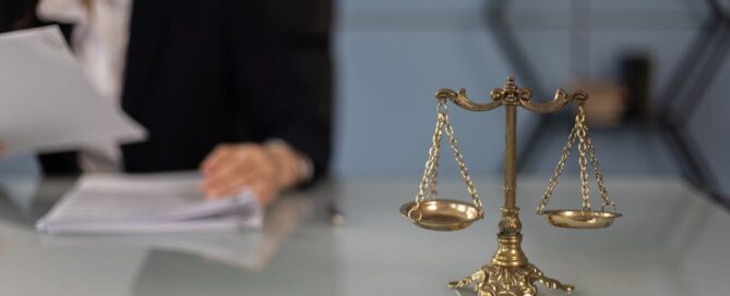 Brass balance scale on a desk with a blurred businesswoman reviewing documents in the background.