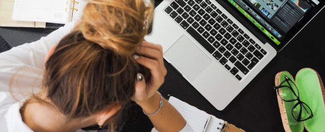Frustrated woman sitting at a desk with a laptop, notebooks, and phone, holding her head in her hands—surrounded by work and paperwork.