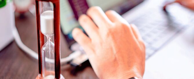 Close-up of an hourglass on a desk beside a person typing on a laptop, symbolising urgency and fast decision-making for business tasks.
