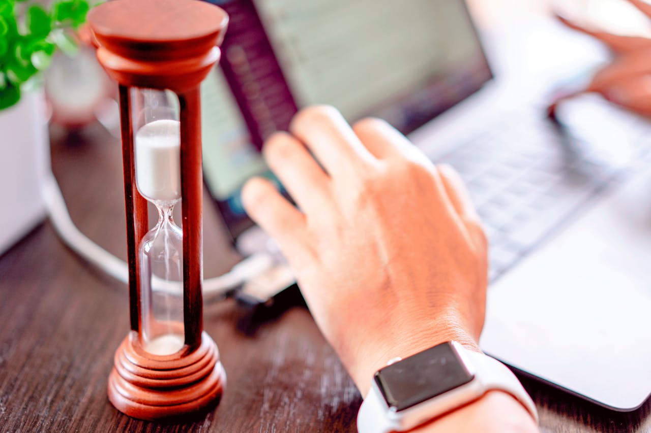 Close-up of an hourglass on a desk beside a person typing on a laptop, symbolising urgency and fast decision-making for business tasks.